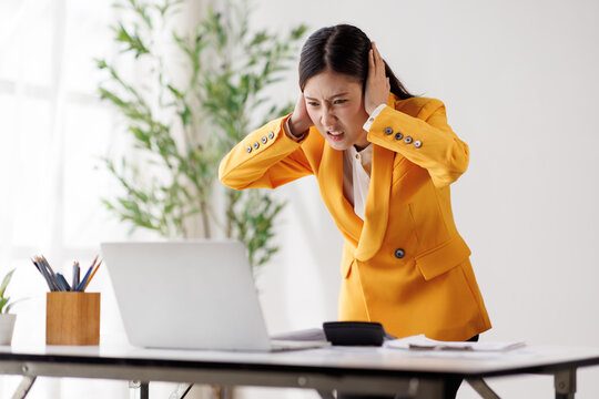 Asian businesswoman covering ears with hands, feeling loud noise, having a headache, stressed by work pressure, experiencing frustration and burnout in the office