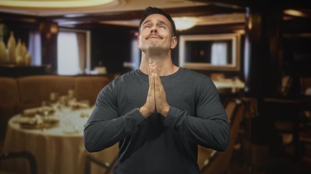 Man with moustache pressing hands together in prayer at a restaurant table inside a building; hopeful plea.