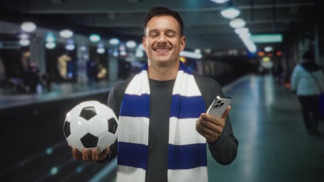Man with moustache holding soccer ball and smartphone, palm presenting ball, wearing striped scarf in subway station; matchday excitement.