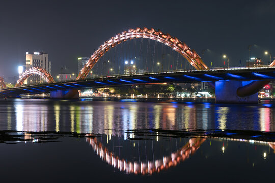 A long Dragon bridge with multiple glowing arches illuminated at night, casting vibrant colorful reflections onto a river in Vietnam