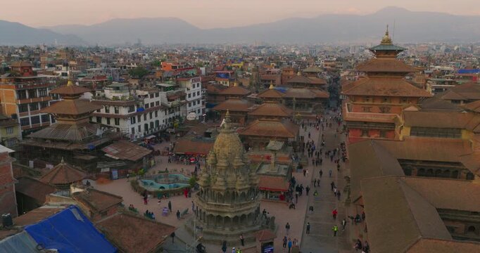 Drone shot of Patan Durbar Square with Krishna Mandir, pagoda-style temples, traditional Newari rooftops, evening visitors and Kathmandu Valley cityscape against hazy hills at sunset.