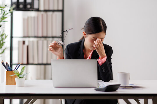 Exhausted businesswoman working at an office desk, experiencing stress, migraine, and headache symptoms while holding eyeglasses, symbolizing eyestrain and burnout from laptop use