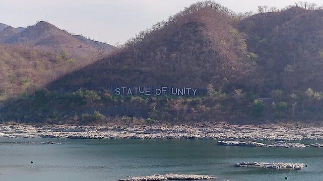 Wide shot of Statue of Unity hillside signage with Narmada River foreground, Ekta Nagar, Kevadia. Ideal for travel docs, India tourism, news, and heritage content.