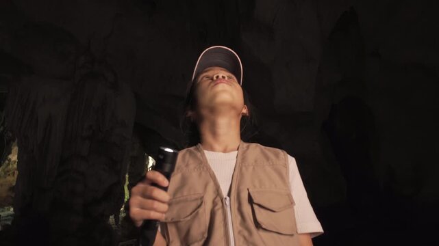 Happy little Asian girl explorer holding a flashlight and looking up inside a dark cave.