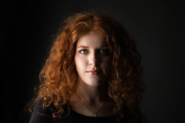 Fototapeta premium Young woman with intense blue eyes and striking red curly hair in dramatic low key lighting