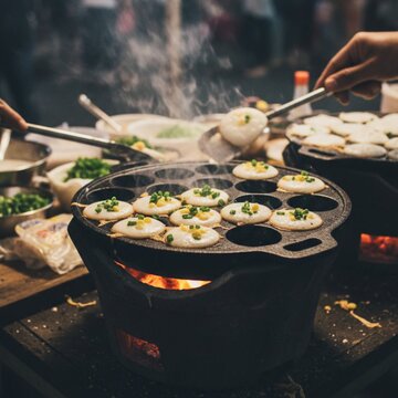 Thai coconut pancakes (Khanom Krok) are being cooked on a traditional cast-iron pan, resting over a live-fire charcoal stove. The pancakes are small, round, and topped with green onions and corn.
