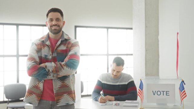 Man with arms crossed and a smiling beard stands by transparent ballot box with american flags at a voting building; civic duty pride.