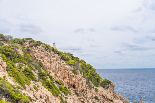 Scenic view of the Capdepera Lighthouse perched on a rocky cliff in Mallorca, Spain, overlooking the ocean beneath a cloudy sky the rugged coastline and distant horizon form.