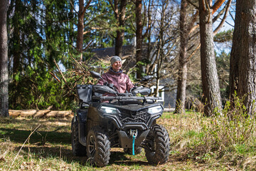 Fototapeta premium A smiling woman is riding an ATV through a forest in Estonia, enjoying the outdoor adventure and exploring the woodland. The all-terrain vehicle is carrying branches for yard.