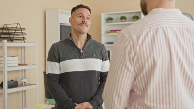 Man in striped sweater gesturing with hand while speaking to coworker in building office; friendly rapport.