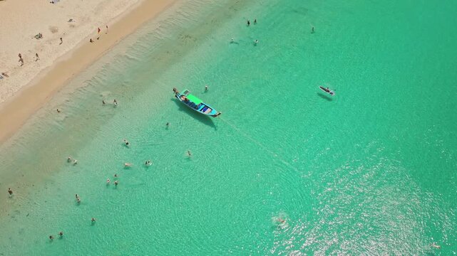 Tropical beach in Phuket, Thailand, with clear turquoise ocean water, a traditional longtail boat anchored offshore, and tourists enjoying swimming and sunbathing under bright sunshine. Aerial view