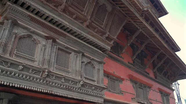 A static low-angle medium shot showcasing rows of intricately carved wooden windows and ancient Newar architecture on a red brick palace wall in Kathmandu, Nepal.