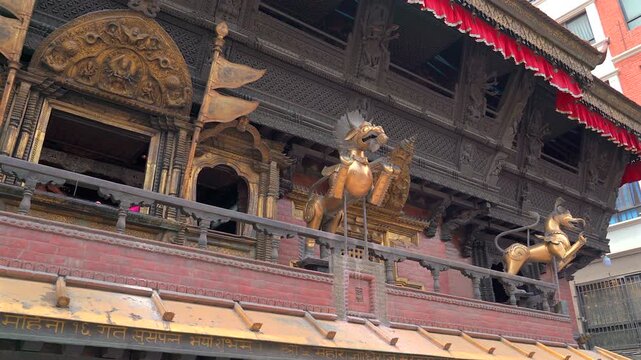 A medium low-angle shot with a subtle pan left revealing the ornate golden mythical griffin statues and intricate dark wood carvings of Akash Bhairav Temple in Nepal.