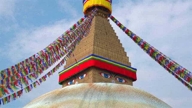 A static medium low-angle shot focusing on the golden spire and sacred wisdom eyes of the Boudhanath Stupa, surrounded by colorful waving prayer flags in Kathmandu, Nepal.