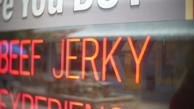 Sign glowing Beef Jerky in a storefront window with soft reflections. Close-up view shows red neon lettering, blurred shop interior, and retail food display
