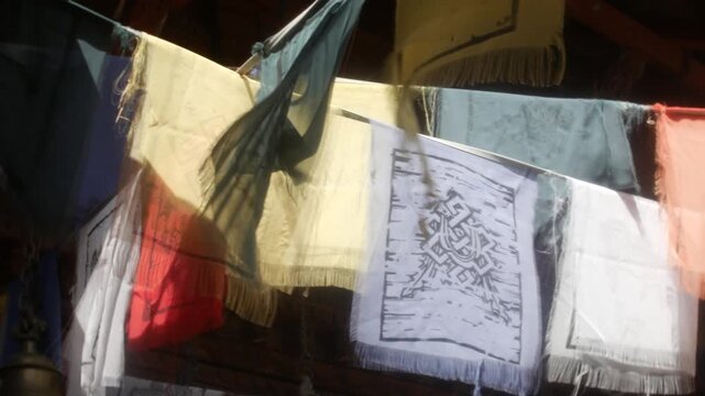 Prayer flags fluttering in soft motion blur beneath dark wooden temple eaves. Close-up view shows patterned fabric, hanging chains, warm shadows, and cultural decor.