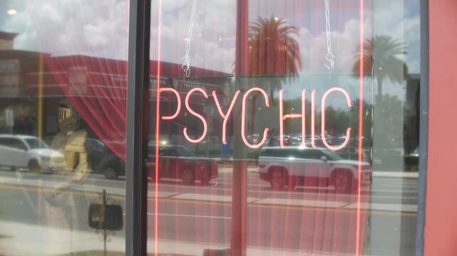 Psychic neon sign glowing in a storefront window with street reflections. Cars, palm trees, red curtains, and bright daylight create an urban retail scene.