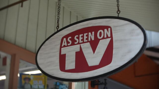 Sign reading As Seen On TV hanging inside a shop near windows and ceiling panels. Close-up view shows red and white lettering on an oval retail display.