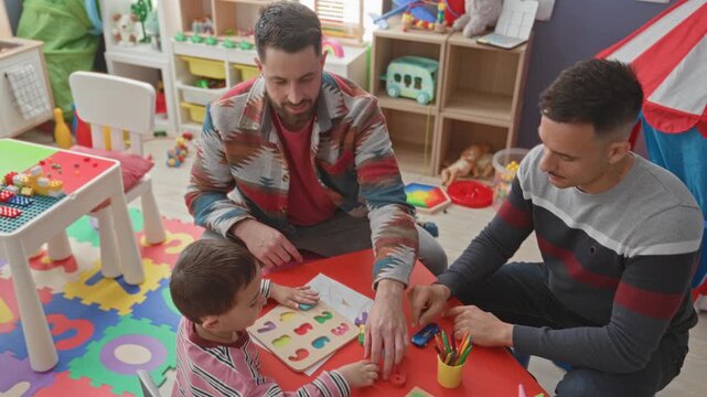 Young boy places wooden puzzle pieces while two men guide with hands at a colorful play table in a building; warmth bonding.
