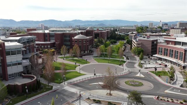 University campus with red brick buildings, circular plaza, roundabout and tree-lined pathways spreading toward downtown skyline and mountain range under partly sunny sky. Nevada, USA. Aerial approach