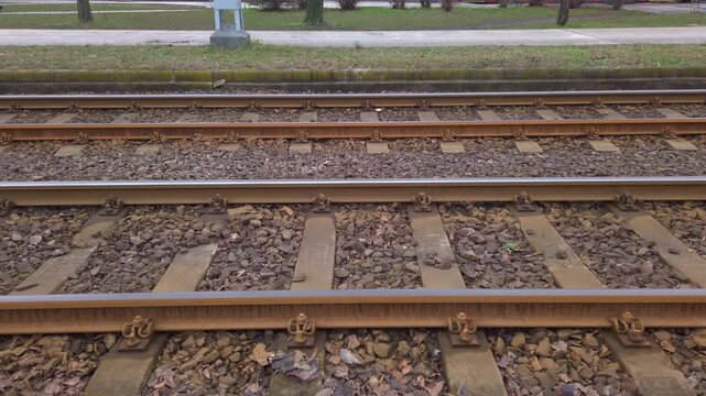 Handheld first person tracking shot moves horizontally along empty parallel steel tram tracks with stone ballast and concrete ties by an urban sidewalk path on a cloudy winter day in Budapest.