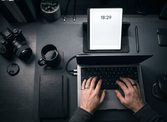 Fototapeta premium Top down view of male hands typing on laptop keyboard on a dark moody workspace with digital tablet mockup, camera, notebook, and coffee.