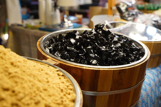 Close up of traditional Thai dessert "Chao Kuay", featuring glossy black grass jelly cubes served in a rustic wooden bucket next to a bowl of brown sugar at a local night market food stall.