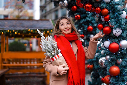 Woman holding a festive arrangement, standing near a decorated Christmas tree in a vibrant outdoor market during the holiday season