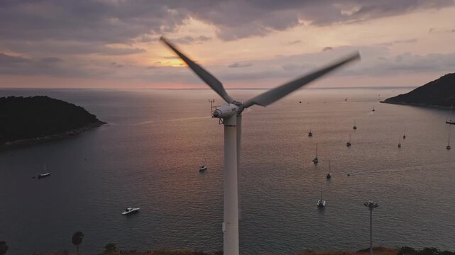 Wind turbine blades spinning. Generating renewable energy on a hillside overlooking a tranquil bay with numerous boats anchored. During a scenic sunset over the Andaman Sea from an aerial perspective