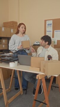 Man and woman pack and scan multiple cardboard parcels at a laptop and handheld scanner in a small shipping area inside a building; focused teamwork.