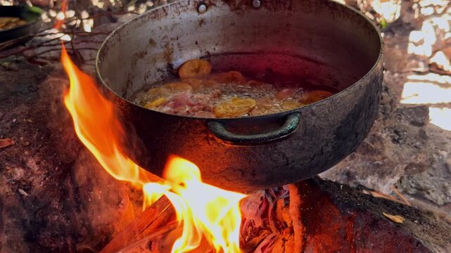 Close-up of chicken pieces and sliced plantains frying in hot oil in large metal pot over open wood fire