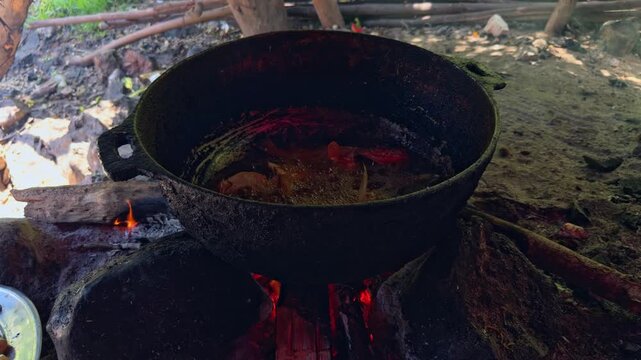 Close-up of whole fish frying in hot oil in large dark metal pot over open wood fire