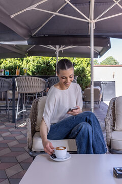 Smiling young woman using her smartphone while reaching for a coffee cup