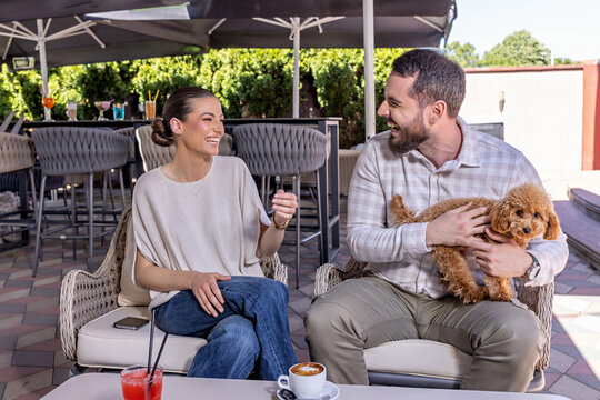 Happy young couple laughing and enjoying coffee at an outdoor cafe terrace with their small puppy