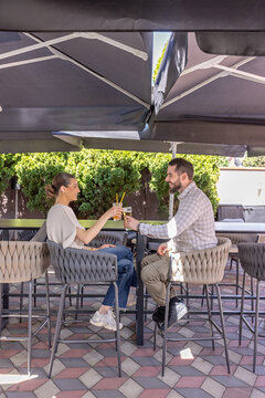 Young man and woman clinking glasses while sitting on high bar stools at an outdoor terrace cafe