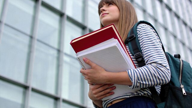Young female student holding stack of notebooks and red book, wearing backpack, standing in front of modern glass university building, education and learning concept.