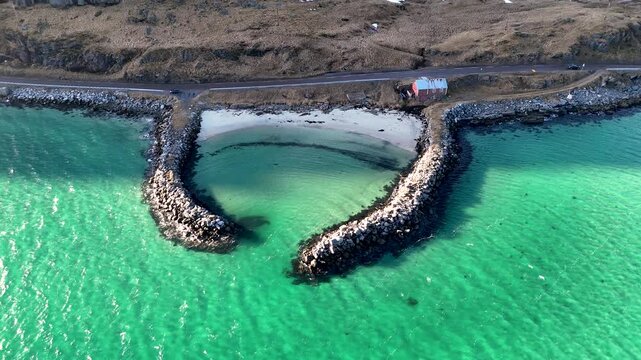 Aerial view of a heart shaped breakwater bay with turquoise water, a red cabin and a scenic coastal road, Lofoten islands Norway, 4K 60fs horizontal Fly Up and Tilt Down drone shot in a sunny day