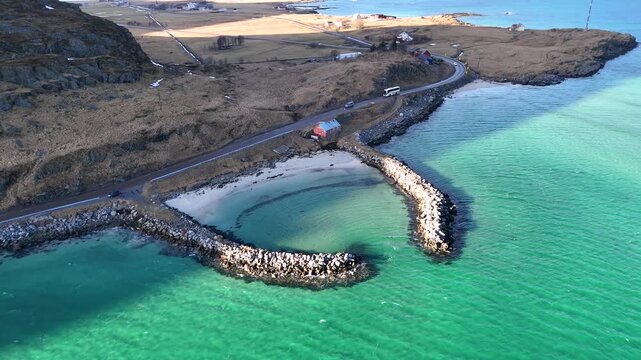Stunning aerial view of a heart shaped breakwater bay with turquoise water, a red cabin and a scenic coastal road with cars, Lofoten islands Norway, 4K 60fs horizontal orbit arc drone shot