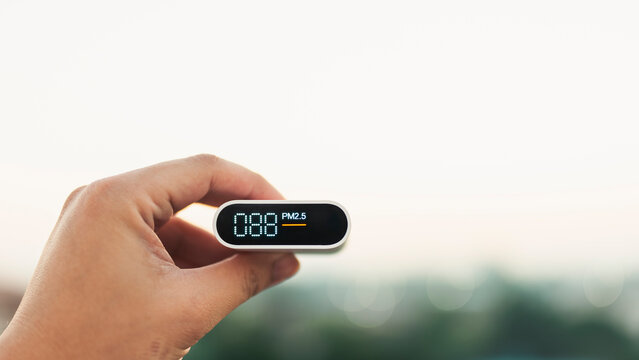 Hand holding a portable air quality monitor showing PM2.5 levels against a blurred outdoor background. Concept of environmental pollution, health care, and smog measurement device.