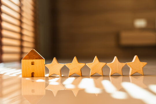 Wooden house model and five stars rating on a reflective surface with bright sunlight. Concept of top quality real estate, premium property, excellent accommodation review, and best service.