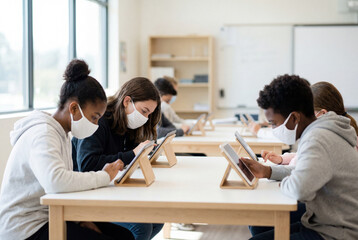 Fototapeta premium Diverse students in masks focused on tablets in modern classroom