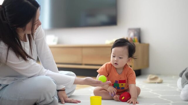 Soft light fills bright, minimalist living room. Mom and baby sit close on patterned floor. Mom gently offers toy to curious infant. Baby reaches with tiny hands, eyes wide with delight