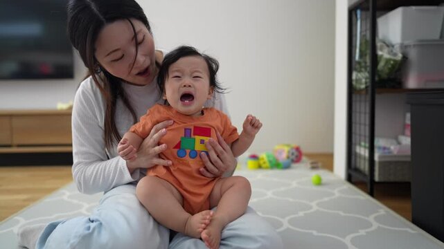 Woman holds laughing baby on floor. Baby cries while mother smiles gently. Toys lie nearby on rug. Home setting feels warm and cozy. Great for parenting, family, or child development themes