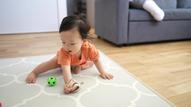 Adorable baby crawls on patterned rug. Curious eyes gaze upward. Tiny hands grasp soft toy nearby. Green ball rests beside them. Cozy living room backdrop. Perfect for parenting, child development