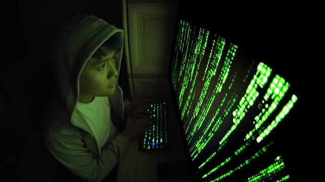 Young person in hoodie types at computer with glowing green code on screen in dark room