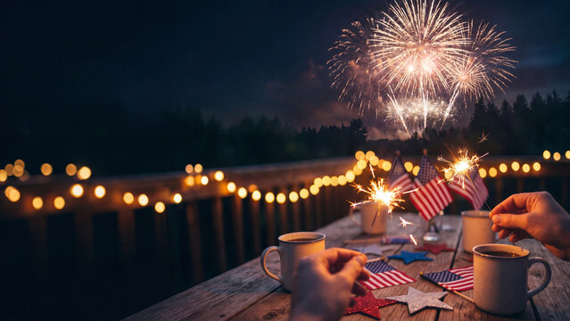 Hands interact with festive American flag decorations and sparklers on a wooden patio table as vibrant fireworks explode in the dark night sky above a suburban home during a summer holiday.