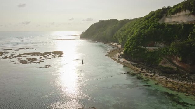 Aerial view Green Bowl Beach in Bali, Indonesia, tropical coastline with lush green cliffs, clear turquoise ocean water, and sunset reflections shimmering over the shallow reefs. Drone flight footage