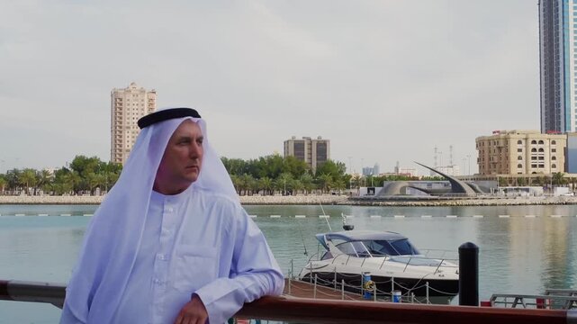 Man in white traditional Gulf Arab thobe and ghutra headwear stands at harbor railing with moored yacht, urban skyline visible across bay with modern buildings.