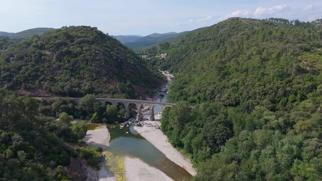 Cinematic drone shot flying forward over an old stone arch bridge spanning across the Gardon de Mialet river. Scenic view deep into the lush green valley in the Cevennes, South of France.