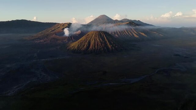 Cinematic drone shot flying backwards over the vast Sea of Sand desert next to green mountain ridges near Mount Bromo during a golden morning sunrise.